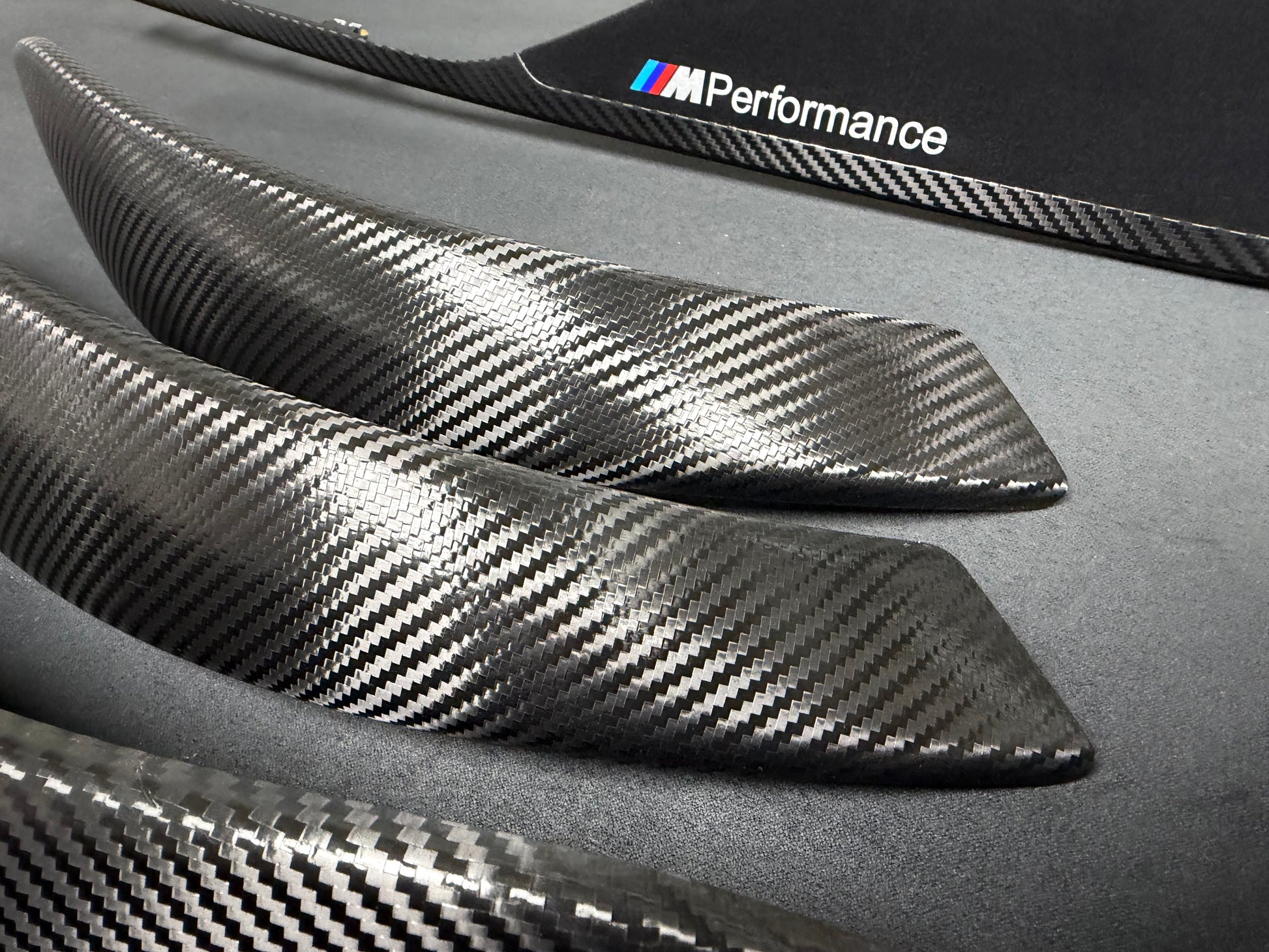 Three carbon fiber trim pieces with a glossy woven pattern, displayed on a dark gray surface. One piece features the white 'M Performance' logo with the classic blue and red stripes.